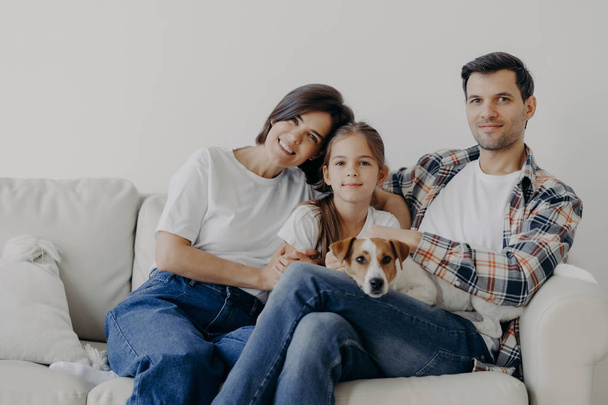Πορτρέτο της στοργικής οικογενειακής αγκαλιάς και κάθονται μαζί στον καναπέ στο σαλόνι, αλλάζουν το σπίτι τους, έχουν χαρούμενες εκφράσεις. Πατέρας, μητέρα, κόρη και σκύλος ποζάρουν για να κάνουν πορτραίτο, περνούν καλά - Φωτογραφία, εικόνα
