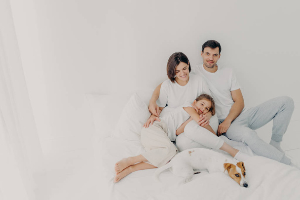 Happy family pose on white bed during weekend. Father, mother, their daughter and dog feel comfort of being in bedroom, embrace and have fun, being relaxed and cheerful. Friendly family and rest - Fotoğraf, Görsel