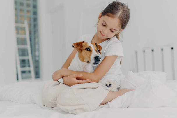 Zdjęcie zrelaksowanej pięknej dziewczynki bawi się z rodowodowym psem, obejmuje ulubione zwierzę, ubrane w luźną piżamę, siedzi na łóżku, wyraża miłość do zwierzaka, pozuje w jasnej sypialni. Szczęśliwego dzieciństwa - Zdjęcie, obraz