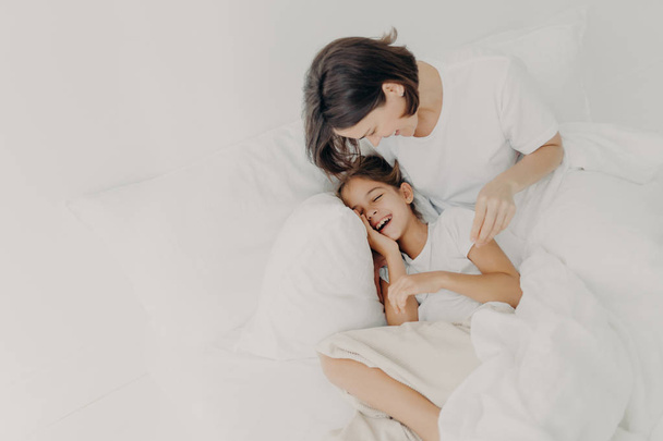 Overjoyed mum and daughter have fun together in bedroom, pose under soft blanket. Happy small girl spends leisure time with mother, has toothy smile, expresses happiness. Children and parents - 写真・画像