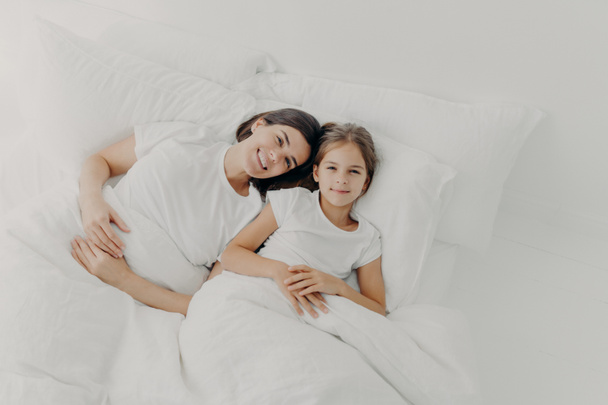 Blick von oben auf glückliche Mutter und Tochter wachen gut gelaunt auf, fühlen sich entspannt, genießen Gemütlichkeit im Bett, freuen sich guten Morgen, im Schlafanzug, liegen unter einer weißen Decke auf weichen Kissen. Bettzeit. - Foto, Bild