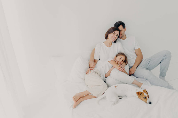 Pleased husband, wife and their small daughter pose in spacious bedroom with favourite pet, have fun, embrace and express love to each other, suppror in difficult situation. Family time concept - Fotografie, Obrázek