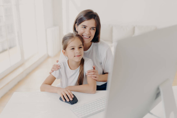 Happy affectionate mom embraces her small daughter, work together on modern computer, preapare presentation for lesson, look gladfully into monitor, spend leisure time at home, surf internet page. - 写真・画像