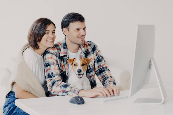 ハッピーフレンドリーなカップルは、犬と一緒に座って、将来の旅行、キーボードのためのホテルの予約をし、コンピュータを見て、オンラインでチャット、白いデスクトップでポーズを取り、無料のwifiに接続し、一緒にインターネットを閲覧 - 写真・画像