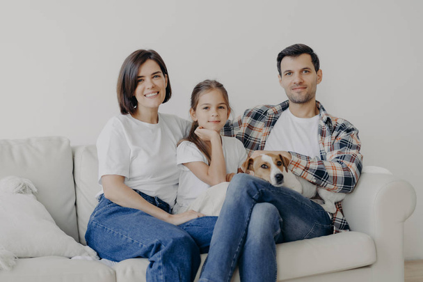 ペットと快適なソファの上でお母さん、お父さんと娘のポーズの幸せな優しい家族の写真は、気軽に服を着て、積極的に笑顔良い関係があります。家庭的な時間と家庭的な雰囲気の概念 - 写真・画像