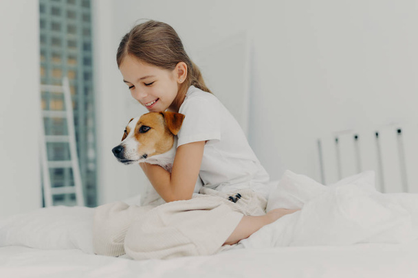 Children, rest and domestic atmosphere. Happy little female child wears nightclothes, cuddle small dog, expresses love and care to favourite pet, pose on comfortable bed with white bedclothes - Photo, Image