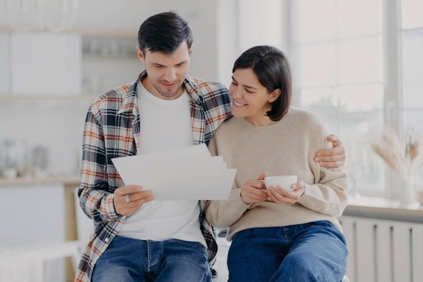 Caring man embraces his wife, look through documents with happy expressions, study payment bills, drinks coffee, dressed in casual wear, pose in spacious room with blurred background. Family and work - Φωτογραφία, εικόνα