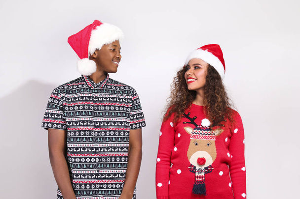 Студийный портрет молодой пары, парня и девушки с темной кожей в шляпе Санта Клауса и рождественском наряде. Уродливая концепция свитера. Закрыть, скопировать пространство для текста, изолированный фон
. - Фото, изображение