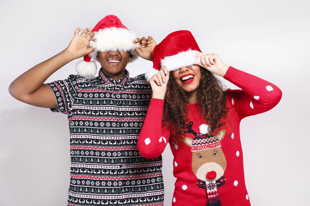 Studio portrait of young couple, boyfriend & girlfriend with dark skin wearing santa claus hat and christmas outfit. Ugly sweater concept. Close up, copy space for text, isolated background. - Fotografie, Obrázek