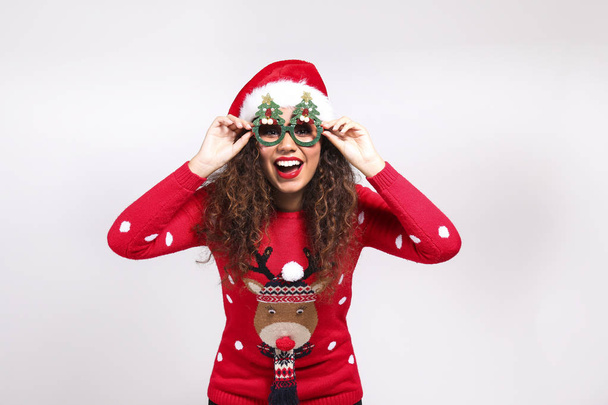 Studio portrait of young woman with dark skin and long curly hair wearing tight santa claus hat and christmas outfit. Ugly sweater concept. Close up, copy space for text, isolated background. - Photo, Image