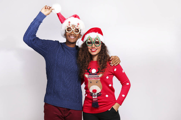 Studio portrait of young couple, boyfriend & girlfriend with dark skin wearing santa claus hat and christmas outfit. Ugly sweater concept. Close up, copy space for text, isolated background. - 写真・画像