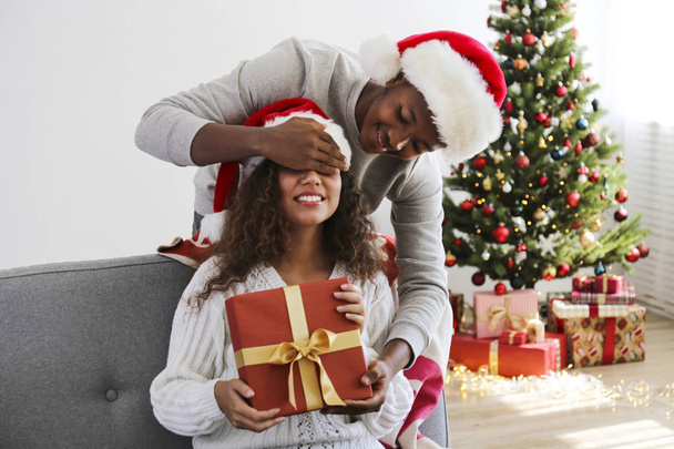 Portrait of young couple, boyfriend & girlfriend with dark skin wearing santa claus hat and christmas outfit exchanging gifts at home. Close up, copy space for text, isolated background. - Fotó, kép