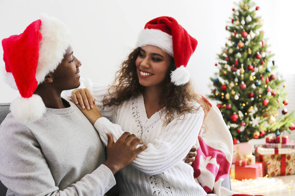 Portrait of young couple, boyfriend & girlfriend with dark skin wearing santa claus hat and christmas outfit exchanging gifts at home. Close up, copy space for text, isolated background. - Fotó, kép