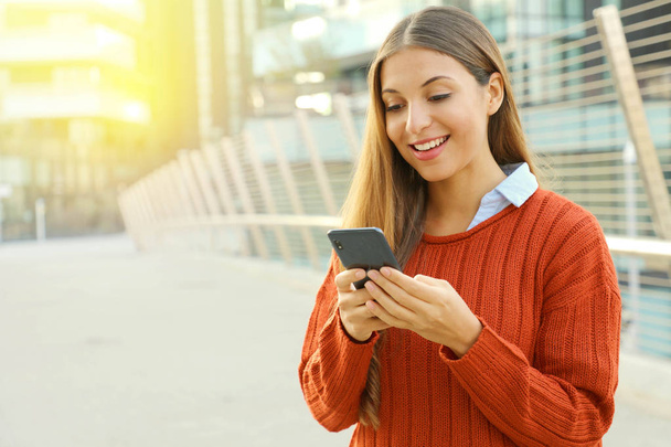 Gelukkige mooie student vrouw schrijven of lezen van sms-berichten online op een smartphone terwijl ze in de moderne stad straat. Kopieerruimte. - Foto, afbeelding