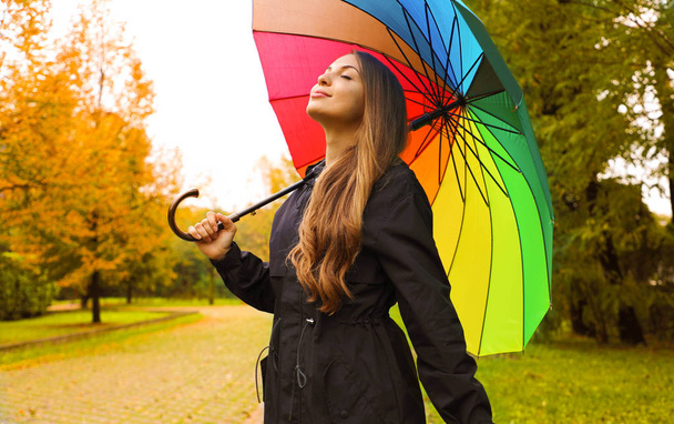 Muotokuva onnellisesta naisesta sadetakissa sateenvarjon alla hengittämässä kaupungin puistossa sadepäivänä
. - Valokuva, kuva