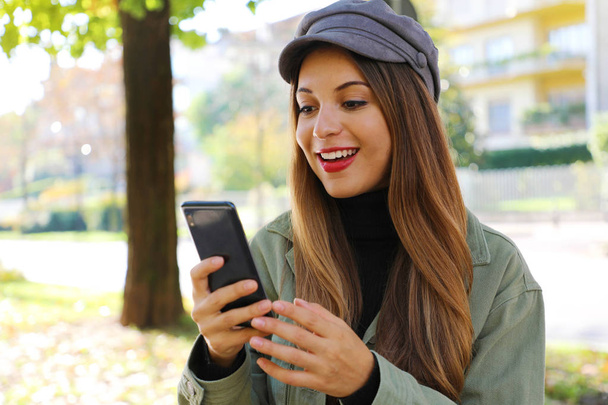 Surprised casual girl wearing hat and polo neck reading good news on smart phone outdoor. - Fotó, kép