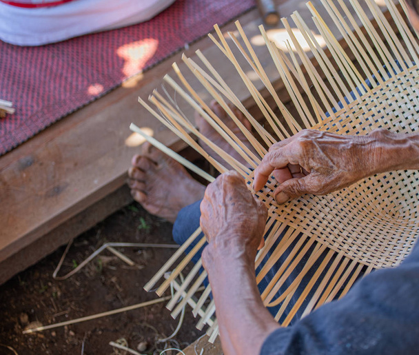 Un habilidoso anciano discapacitado hace mimbre de bambú con las manos en el suelo. Mostrando simplemente el modo de vida mediante la práctica de la autosuficiencia
. - Foto, Imagen