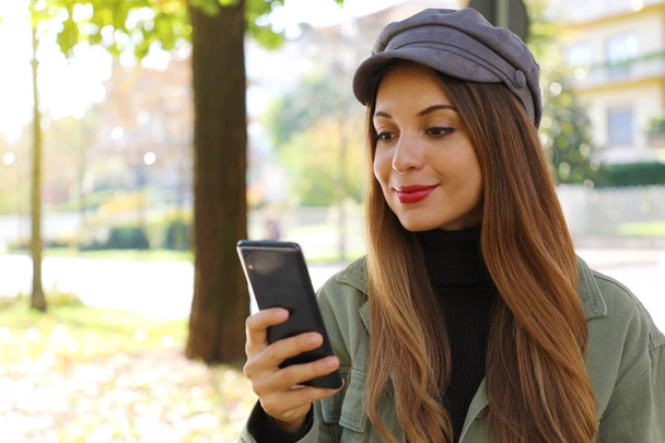 Lässiges Mädchen mit Hut und Polohals tippt auf Smartphone im Freien. - Foto, Bild