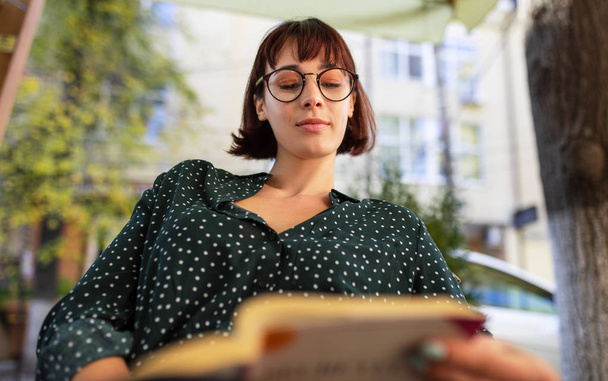 Image of beautiful young woman wearing green shirt with white dots and transparent eyeglasses, sitting outdoor on the city street and reading book. Young female student learning in the city street.  - 写真・画像