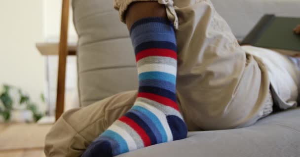 Бічний вид на низьку частину молодого афроамериканського хлопчика, який лежав на дивані в своїй вітальні в смугастих шкарпетках спокійно читає книгу самостійно, повільно рухаючись - Кадри, відео