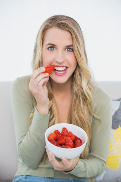 Happy cute blonde eating strawberries sitting on cosy sofa - Photo, Image