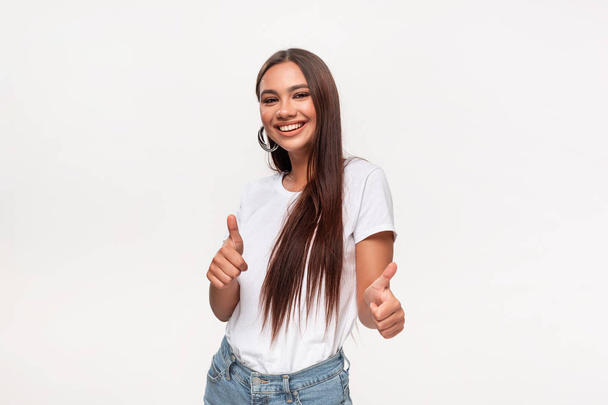 Excited pretty african-american teenager in a white t-shirt and blue jeans holding thumbs up  isolated over white background. - Foto, Imagen