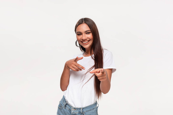 Excited pretty african-american teenager in a white t-shirt and blue jeans holding fingers in a finger gun gesture. - Photo, image