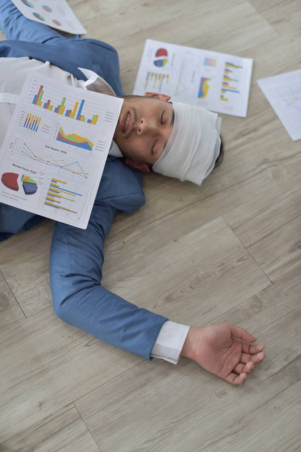 Hombre de negocios sobrecargado de trabajo con la cabeza herida vendada acostado en el suelo con documentos dispersos
 - Foto, imagen