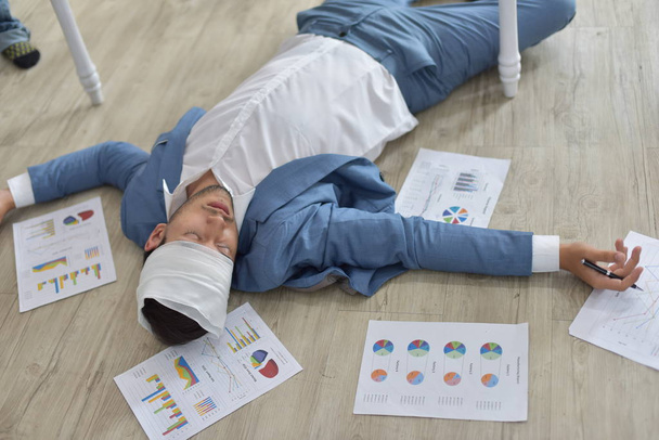 Υπερκόπωση επιχειρηματίας με επίδεσμο χτυπημένο κεφάλι ξαπλωμένος στο πάτωμα με διάσπαρτα έγγραφα - Φωτογραφία, εικόνα