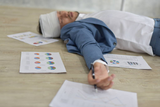 Υπερκόπωση επιχειρηματίας με επίδεσμο χτυπημένο κεφάλι ξαπλωμένος στο πάτωμα με διάσπαρτα έγγραφα - Φωτογραφία, εικόνα