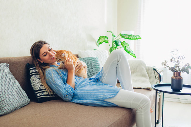 Playing with cat at home. Young woman sitting on couch teasing and hugging pet. - Fotografie, Obrázek