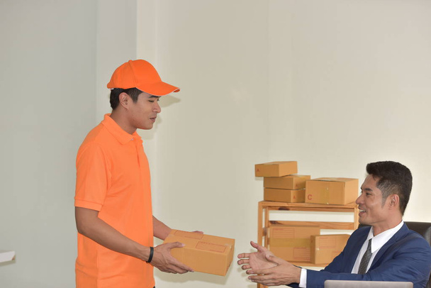 Koncepcja usługi dostawy, młody pracownik azjatycki w pomarańczowym mundurze dostarczanie paczek dla biznesmena. - Zdjęcie, obraz
