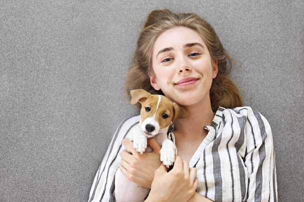 The cutest two months old Jack Russel terrier puppy with beautiful blonde young woman. Small adorable doggy with funny fur stains lying with owner. Close up, copy space, isolated background. - 写真・画像