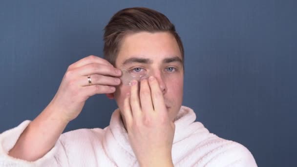 A young man puts patches on his face. Blue hydrogel patches for rejuvenating the skin of the face. Man sitting in a white coat. Close up - Materiaali, video