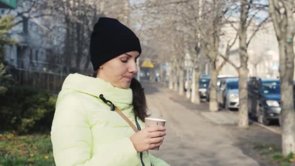girl in a down jacket is walking down the street with a glass of coffee in her hands - Imágenes, Vídeo