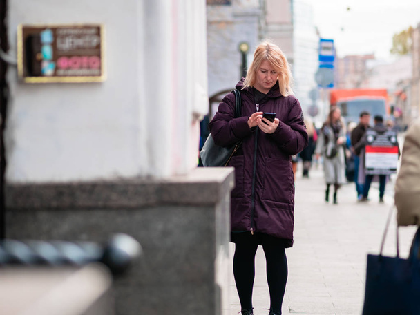 Moscow, Russia - October 19, 2019: A middle-aged woman with blond hair is typing a message on a smartphone. The lady is wearing a purple light overcoat. Warm autumn sunny day - Foto, afbeelding