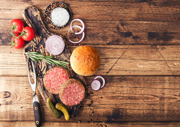 Fresh raw minced pepper beef burgers on vintage chopping board with buns onion and tomatoes on wooden background. Space for text - Foto, Imagem