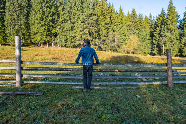 Young male farmer in a denim jacket and cowboy hat stand near the wooden fence against forest  - Foto, imagen