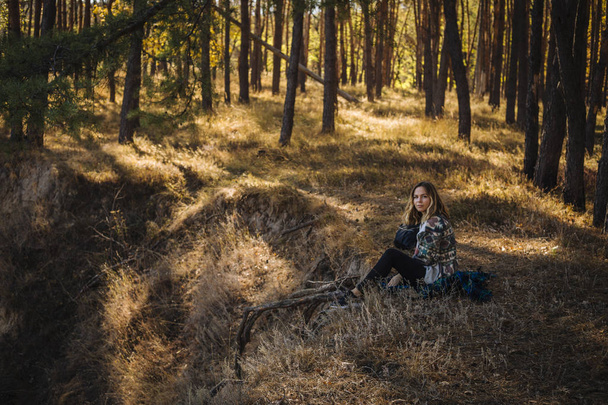 Νεαρή όμορφη ξανθιά κοπέλα με σγουρά μαλλιά κάθεται στο έδαφος σε ήσυχο δάσος φθινόπωρο - ήρεμη και διαλογιστική. - Φωτογραφία, εικόνα