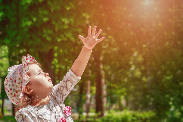 Ένα μικρό κορίτσι στέκεται σε ένα πάρκο απλώνεται στις ακτίνες του ήλιου - Φωτογραφία, εικόνα