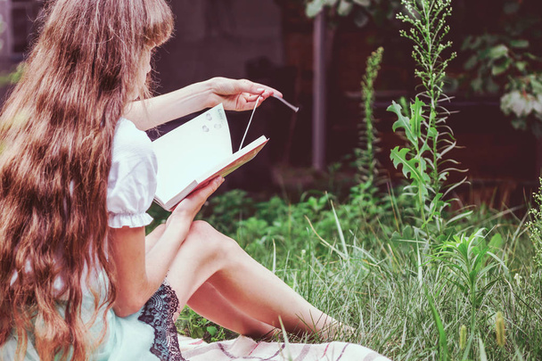 A teenage girl with long blond hair and a backpack is sitting on the grass and reading a personal diary. Concept - secret life, lifestyle - Foto, Bild