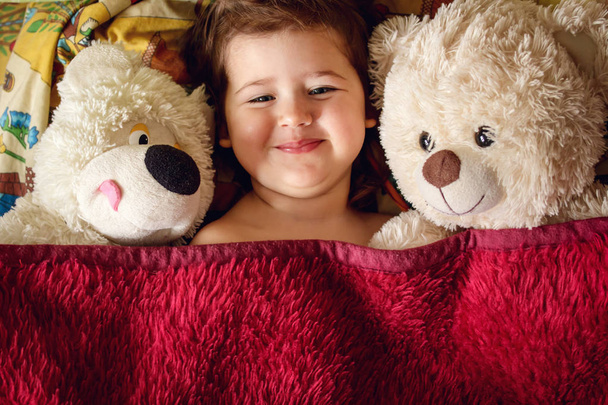 The morning of a small smiling girl with two soft bears under the blanket - Foto, afbeelding