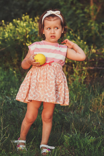 Маленькая девочка в платье с рисунком смотрит в сторону и держит лимон, открытый воздух, крупный план
 - Фото, изображение