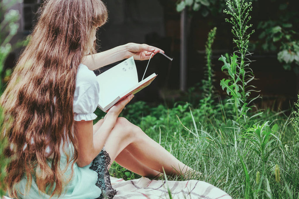 A teenage girl with long blond hair and a backpack is sitting on the grass and reading a personal diary. Concept - secret life, lifestyle - Foto, Imagen