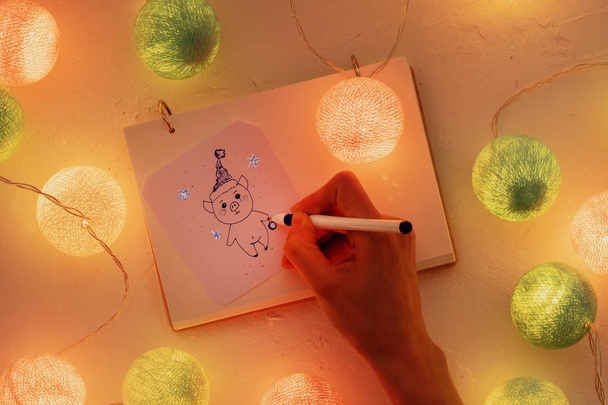 Female hand close-up draws a piglet, around luminous garlands. Concept - New Year's decor, symbols and festive mood - Photo, Image