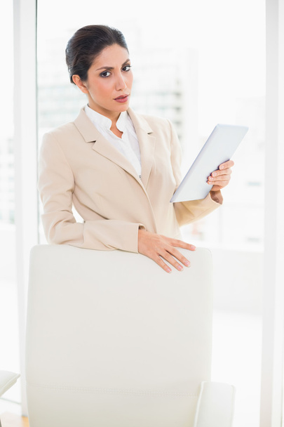 Frowning businesswoman standing behind her chair holding tablet pc - Foto, Imagem