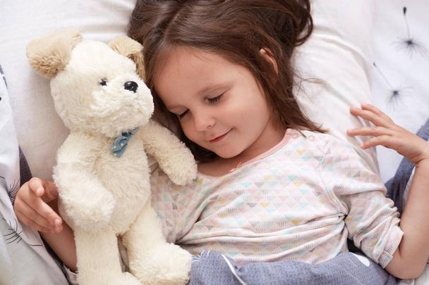 Schattig klein meisje lag met gesloten ogen, kind in witte pyjama liggend op een wit dekbed, slapend in een comfortabele kamer, schattig kind speelt met haar speeltje en valt in slaap. Het begrip "jeugd". - Foto, afbeelding