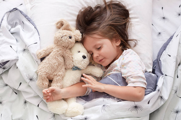 Sweet little girl sleeping with toys in crib. Close up portrait of infant sleeping in cot. Beautiful toddler sleeping with toy bear and dog, tired baby relax. Sweet bedtime and childhood concept.. - Foto, Imagen