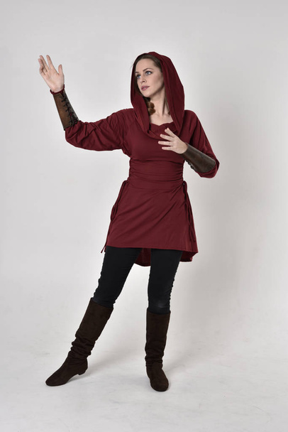 pełnowymiarowy portret brunetki ubranej w czerwoną tunikę fantasy z kapturem. Pozycja stojąca na białym tle studia. - Zdjęcie, obraz