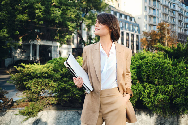 Молодая привлекательная улыбающаяся деловая женщина в бежевом костюме стоит с ноутбуком на городской улице
 - Фото, изображение